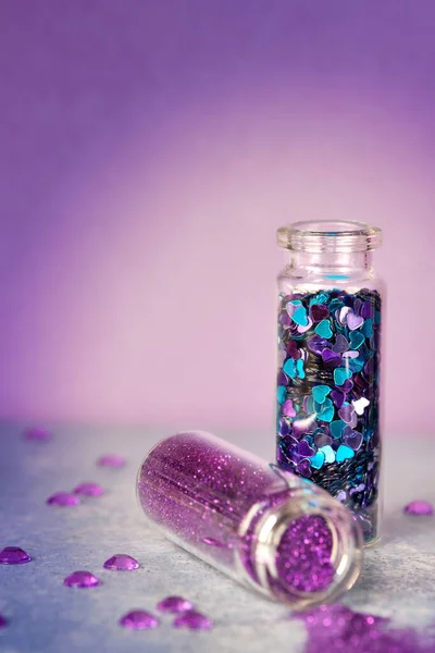 All kinds of glitter products on pink sparkling background. Close-up on vials, bottles with various glitter makeup in pink, blue and turquoise shades. Shallow DOF, focus on scattered violet glitter.