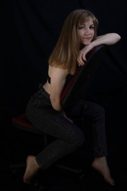 İnce vücutlu, seksi ve güzel bir sarışın. Uzun baştan çıkarıcı bacakları var. Sandalyede kot pantolon, arka planda koyu renk bir duvar var.