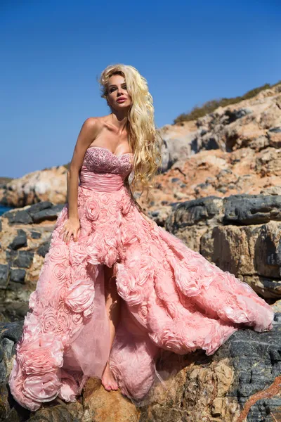 Hermosa mujer rubia sexy vestido de salón rosa de pie sobre las rocas en Santorini — Foto de Stock