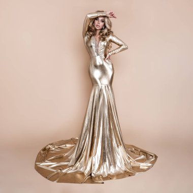 Zarif bir moda. Zarif, uzun, altın sarısı elbisesi ve bej arka planda mükemmel saç stili olan sarışın kadın. Lüks akşam modası. Cazibeli manken. Zarafet. Altın elbise.