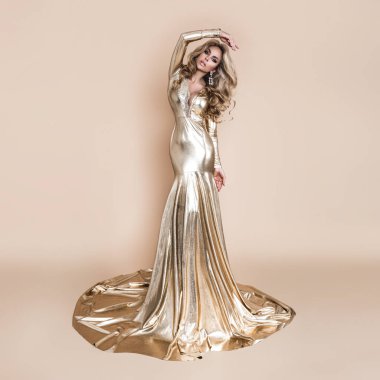 Zarif bir moda. Zarif, uzun, altın sarısı elbisesi ve bej arka planda mükemmel saç stili olan sarışın kadın. Lüks akşam modası. Cazibeli manken. Zarafet. Altın elbise.