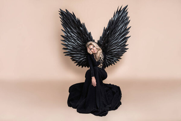Elegant fashion. Stunning blonde woman in elegant long black dress and big wings on beige background in studio. Luxury evening fashion. Glamour fashion model. Classy woman. Woman with wings. Angel.