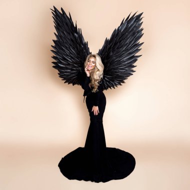 Zarif bir moda. Zarif, uzun, siyah elbiseli ve bej arka planda büyük kanatlı sarışın kadın stüdyoda. Lüks akşam modası. Cazibeli manken. Çok klas bir kadın. Kanatlı kadın. Melek.