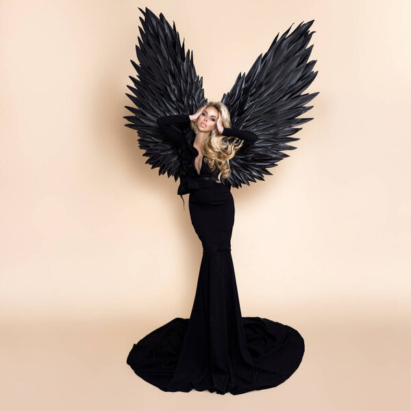 Elegant fashion. Stunning blonde woman in elegant long black dress and big wings on beige background in studio. Luxury evening fashion. Glamour fashion model. Classy woman. Woman with wings. Angel.