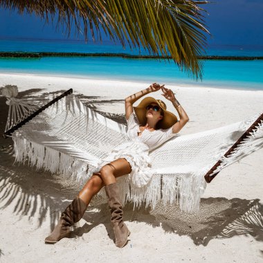 Şık elbiseli zarif bir kadın ve Maldivler adasındaki tropik plajda büyük bir şapka. Yaz modası. Kadın model kumlu sahilde poz veriyor. Maldivler manzarası. Lüks Seyahat.