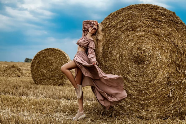Beautiful female model in a fashionable dress and shoes is posing in a grain field on summer day. Sensual woman in country style posing against the countryside. Outdoor rural session. Rural girl.