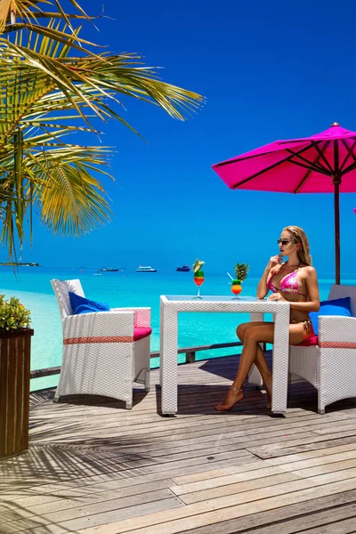 Beautiful woman in bikini is sitting in beach bar on the Maldives sandy beach and holding colorful fruity drink. Luxury travel. Tourism. Maldives landscape.