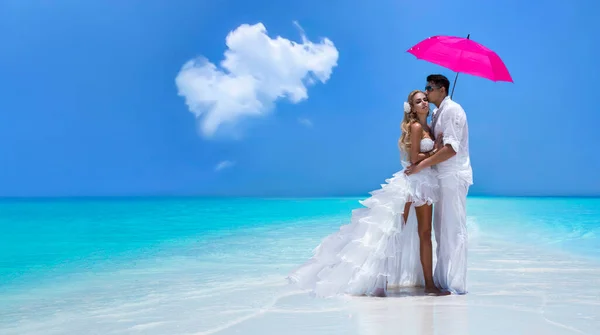 Summer love. Beautiful happy young couple in wedding clothes and pink umbrella is standing on a beach in the Maldives. Engagements and wedding on the beach on the paradise island of Maldives. Luxury travel.