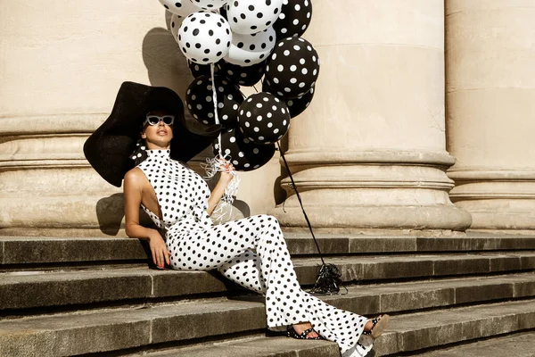 Spring, summer fashion. Glamour, stylish elegant woman in polka dot jumpsuit is holding balloons with dots. Fashion model in outfit with polka dots in the city. 60's style. Retro fashion
