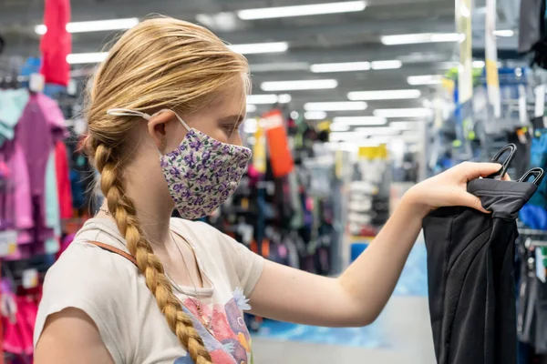 New normal retail shopping. Teenage blond girl wearing face mask choosing sports clothing at sport warehouse retail shop. Covid-19 pandemic