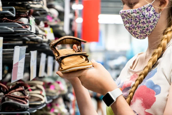 New normal retail shopping. Teenage blond girl wearing face mask choosing walking shoes sandalds at sport warehouse retail shop. Covid-19 pandemic