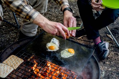 Kahvaltı kampı yemeği. Adam kamp ateşinin üzerinde yumurta pişiriyor ve demir bir tabakta ekmek ısıtıyor. Kamp yaşam tarzı