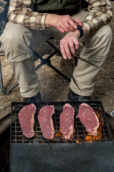 Man cooking beef steaks on a cast iron grlill plate on a camp fireCampfire camping cooking. Outdoor BBQ