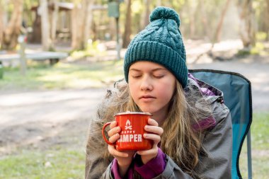 Kamp yaşam tarzı. Kamp ateşinin yanındaki kamp sandalyesinde oturan bereli genç bir kız elinde kırmızı bir diş minesi bardağı tutuyor. Mutlu kampçı.