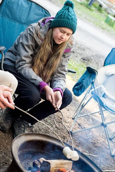 Teenage girl wearing beanie hat roasting large marshmallow on a stick over the campfire firepit. Camping family fun lifestyle