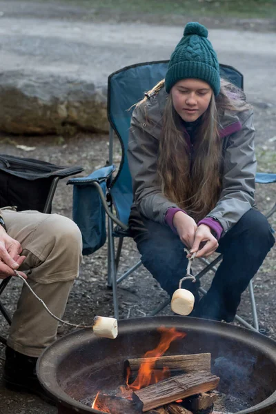 Teenage girl wearing beanie hat roasting large marshmallow on a stick over the campfire firepit. Camping family fun lifestyle