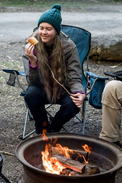 Teenage girl wearing beanie hat eating large marshmallow on a stick roasted over the campfire firepit. Camping family fun lifestyle