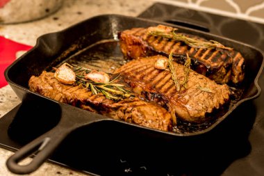 Grilling cooking New York beef sirloin strip steaks with rosemary and garlic on cast iron grill pan skillet on a ceramic stove counter top at home.