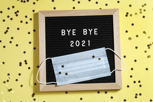 Tablero de letras negro con texto BYE BYE 2021 con pequeñas estrellas y máscara sobre fondo amarillo. — Foto de Stock