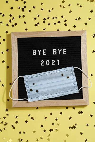 Tablero de letras negro con texto BYE BYE 2021 con pequeñas estrellas y máscara sobre fondo amarillo. — Foto de Stock