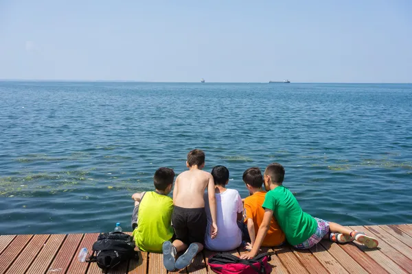 Kids sitting on wooden platform beside polluted sea with clear s — Stock Photo, Image