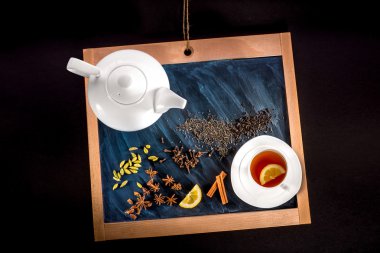cup of tea and teapot with spices and lemon on chalkboard on bla