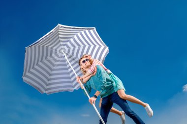 Girl having piggy back ride on a man with beach umbrella on blue clipart