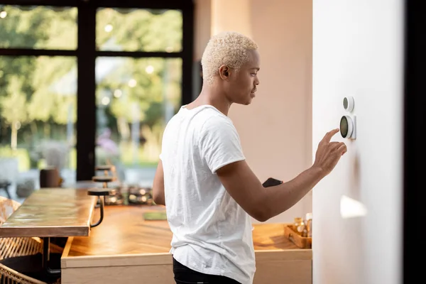 拉丁男人选择恒温器上的温度 年轻专注的家伙按下了智能家庭系统的按钮 现代家庭生活方式的概念 现代公寓厨房的内部 — 图库照片