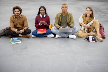 Dijital aygıtları olan öğrenciler asfaltta oturuyorlar