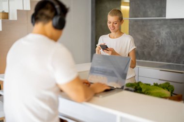 Kadın yemek pişirirken, erkek bilgisayar kullanıyor.