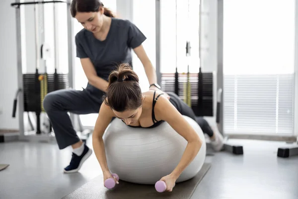 Vrouw doet oefeningen op fitness bal en halters met revalidatie specialist in de sportschool — Stockfoto