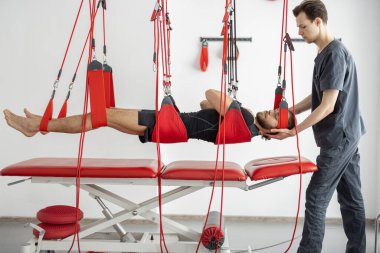 Rehabilitasyon uzmanı erkek hastaya süspansiyon kayışları üzerinde aktif tedavi uyguluyor.