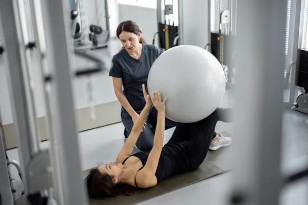 Γυναίκα κάνει ασκήσεις με μπάλα γυμναστικής με ειδικό αποκατάστασης στο γυμναστήριο — Φωτογραφία Αρχείου