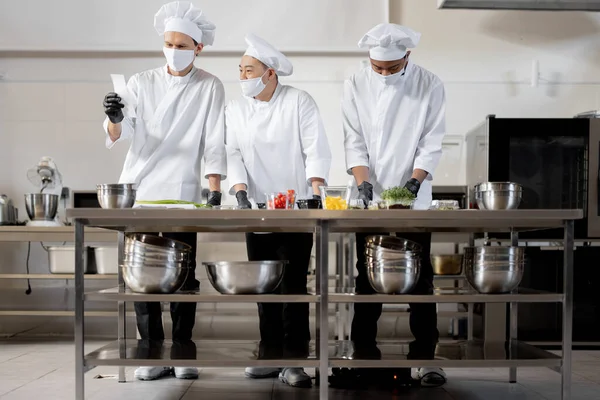 Chefs en mascarillas preparan comida para llevar en cocina profesional — Foto de Stock