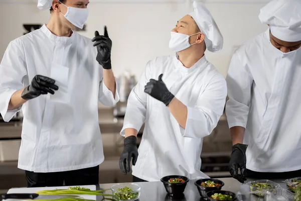 Chefs em máscaras faciais preparar comida takeaway na cozinha profissional — Fotografia de Stock