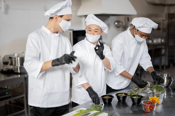 Chefs en mascarillas preparan comida para llevar en cocina profesional —  Fotos de Stock