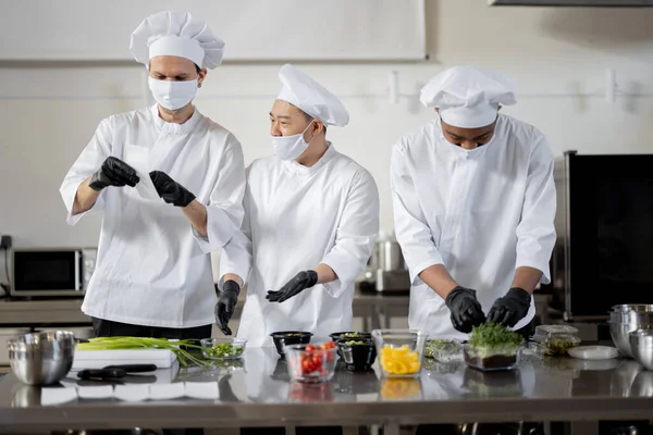 Chefs em máscaras faciais preparar comida takeaway na cozinha profissional — Fotografia de Stock