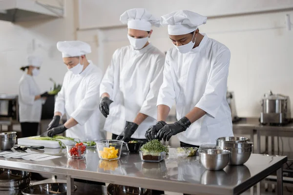Chefs en mascarillas preparan comida para llevar en cocina profesional —  Fotos de Stock