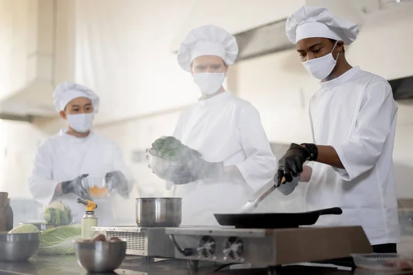 Équipe multiraciale de cuisiniers en uniforme et masques faciaux cuisiner les repas pour un restaurant dans la cuisine — Photo