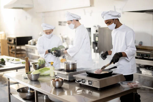 Equipo multirracial de cocineros en uniforme y mascarillas para cocinar comidas para un restaurante en la cocina —  Fotos de Stock