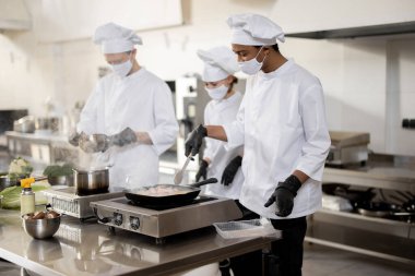 Üniformalı ve maskeli aşçılardan oluşan çok ırklı bir ekip mutfaktaki bir restoranda yemek pişiriyor.