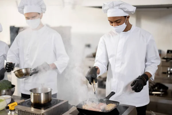 Dois chefs de uniforme e máscaras faciais cozinhando juntos na cozinha profissional — Fotografia de Stock