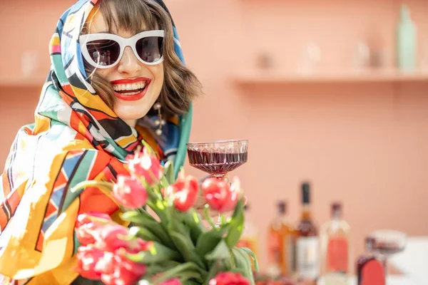Traço de uma mulher elegante em xale colorido e óculos de sol com flores e bebida comemorando dentro de casa — Fotografia de Stock