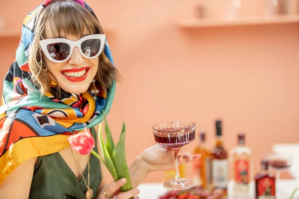Rtrait av en elegant kvinna i färgglada sjal och solglasögon med blommor och dryck firar inomhus — Stockfoto