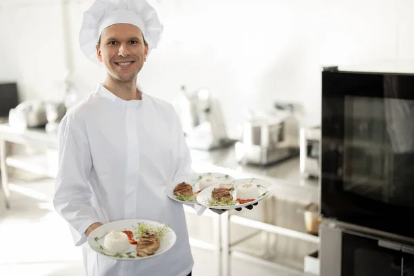 Cuoco sorridente in uniforme in piedi con piatti pronti in cucina — Foto Stock