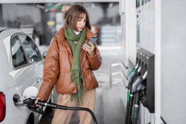 Yüz maskeli kadın akıllı telefon kullanarak arabaya benzin dolduruyor.