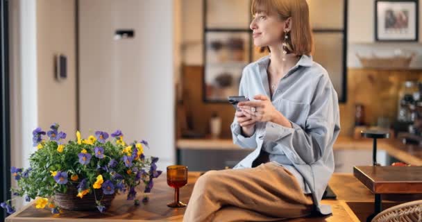 La mujer se sienta en la parte superior de la cocina con teléfono en la casa hermosa — Vídeos de Stock