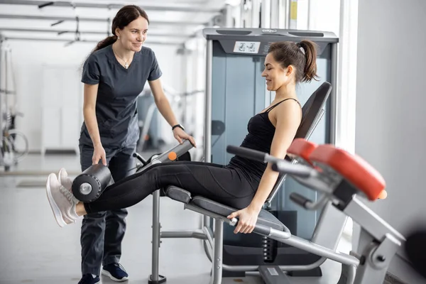 Kobieta robi ćwiczenia na symulatorze ze specjalistą rehabilitacji na siłowni — Zdjęcie stockowe
