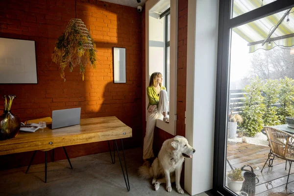 Nainen istuu ikkunalaudalla ja katselee ikkunasta valkoisen hurmaavan koiransa kanssa kotona. — kuvapankkivalokuva