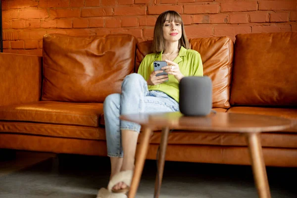 Kvinna med telefon och smart högtalare sitter avslappnad hemma — Stockfoto